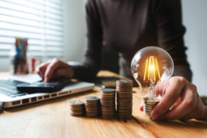 A woman uses a calculator at her desk while looking at a lightbulb and a pile of coins.