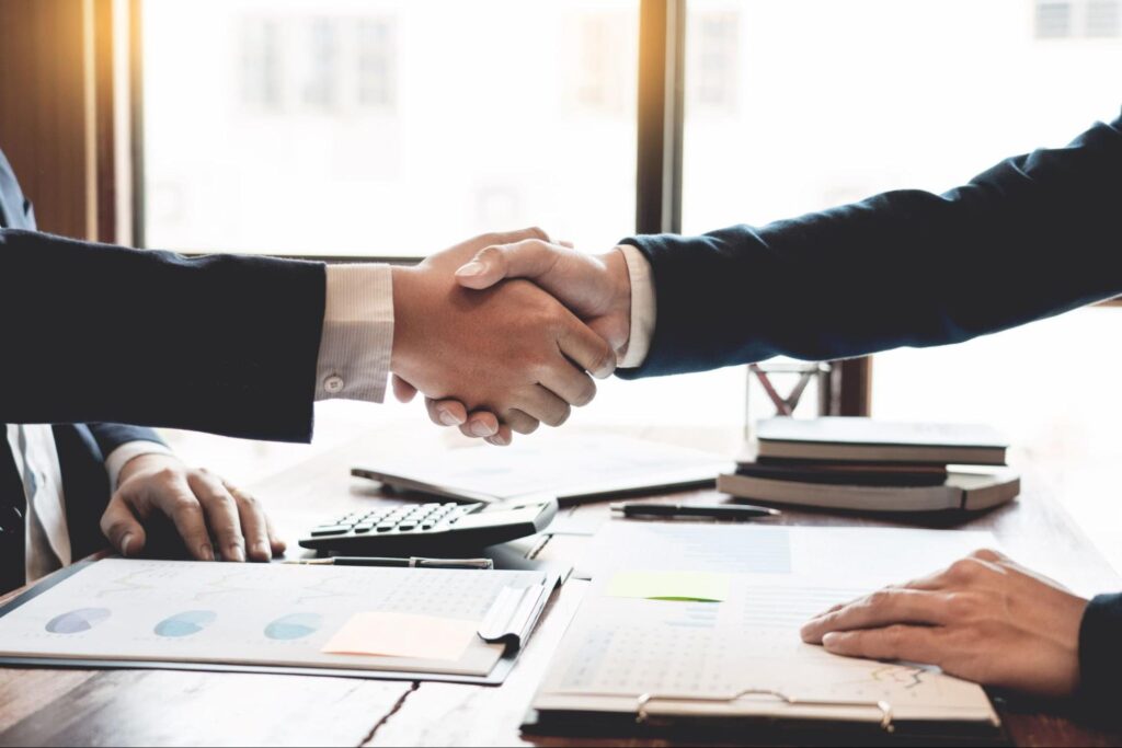 Two people shake hands over a desk with files of data and graphs in front of them.