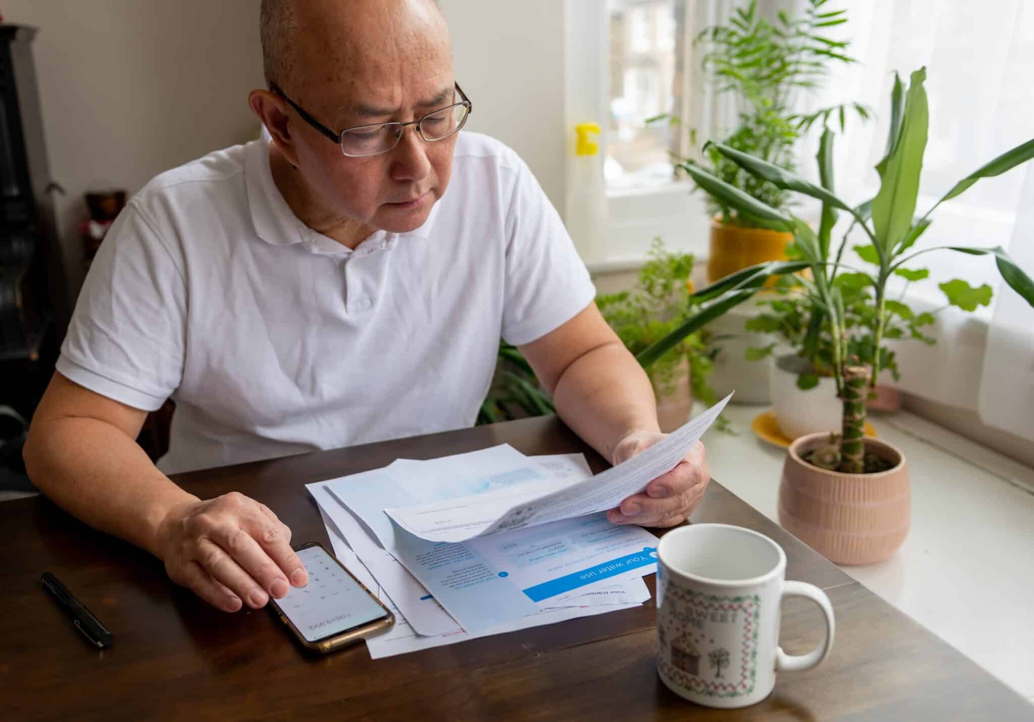 A man peruses his energy bill at a desk using a mobile phone as a calculator.