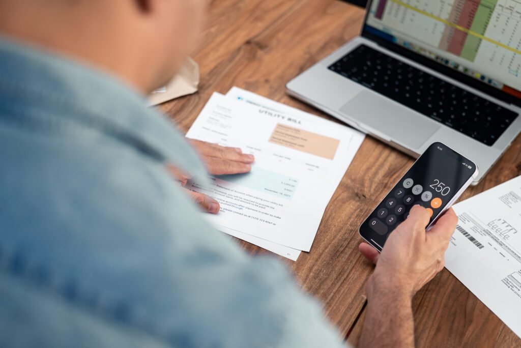 Man using a smartphone calculator to review utility bills at a wooden table. Financial planning and household budgeting concept. Ideal for expense management and personal finance topics